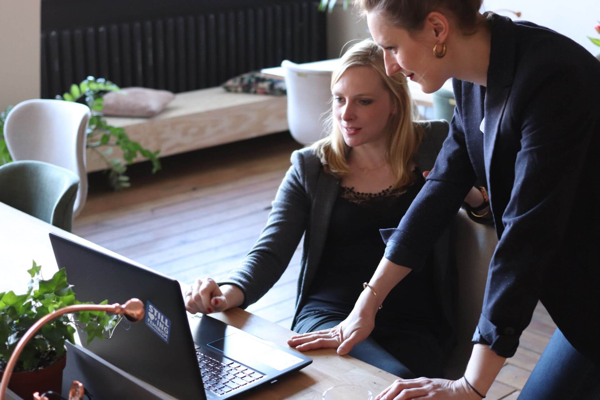 photo of 1 woman sitting and another standing, looking at laptop on desk